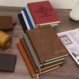 May include: A collection of personalized journals in various colors, including brown, gray, and burgundy. The burgundy journal features the initials "H M" with floral accents and the names "Hailey Megan." The brown journal is inscribed with the name "Amelia."