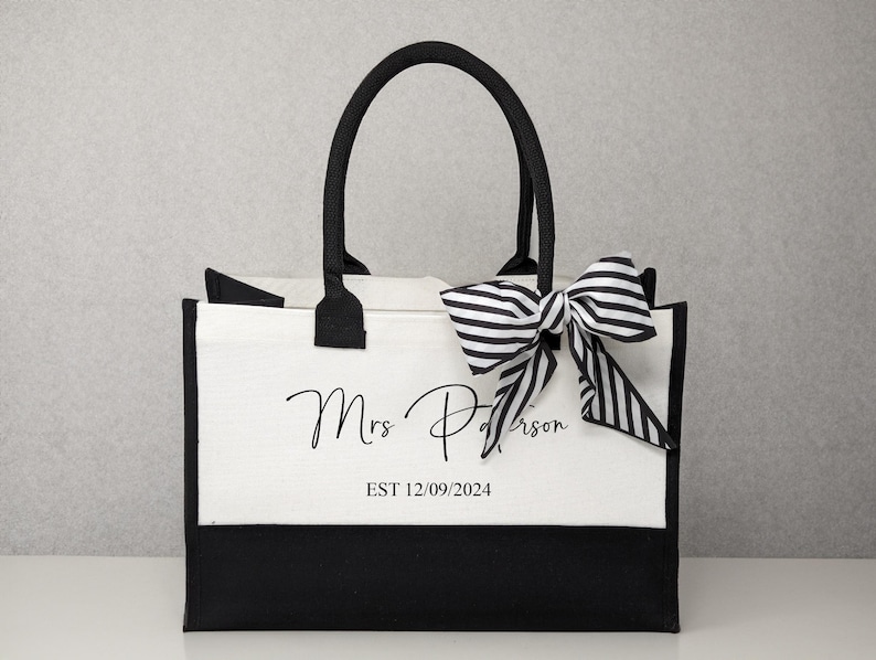 K&ouml;nnte beinhalten: Eine wei&szlig;e und schwarze Tragetasche mit schwarzen Henkeln. Die Tasche hat eine schwarz-wei&szlig; gestreifte Schleife oben und den Text "Mrs. Patterson EST 12.09.2024" auf der Vorderseite.