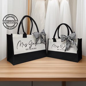 K&ouml;nnte beinhalten: Zwei schwarze und wei&szlig;e Tragetaschen mit schwarzen Griffen und schwarz-wei&szlig;en Schleifen. Die Taschen sind mit dem Text "Mrs. Garcia EST. 05.26.2024" und "Mrs. Wilson EST. 10.10.20" personalisiert.