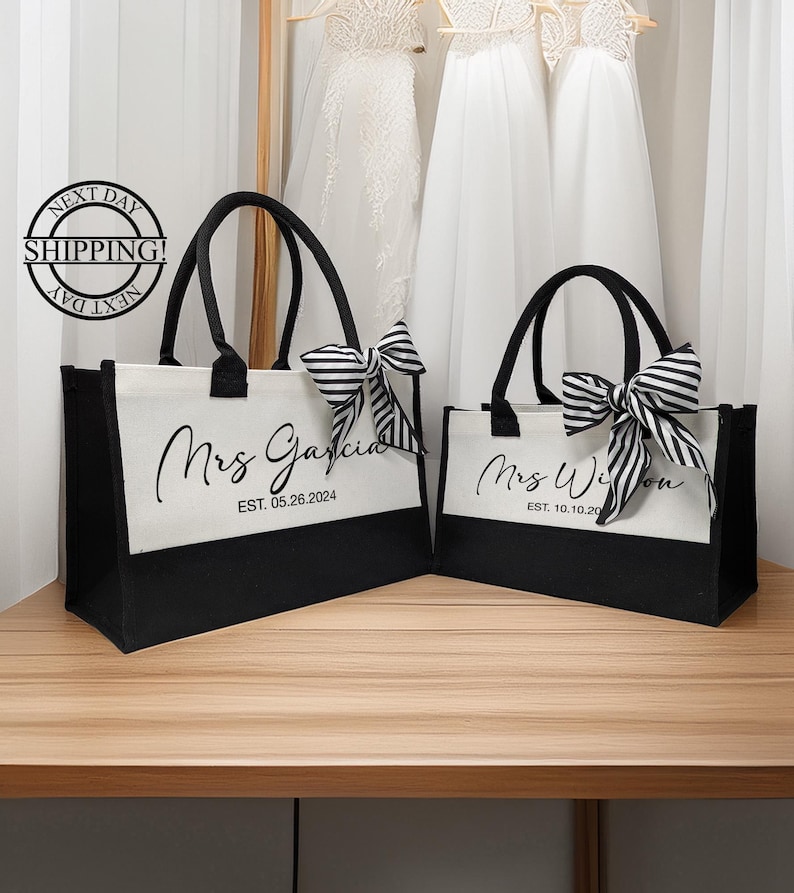 K&ouml;nnte beinhalten: Zwei schwarze und wei&szlig;e Tragetaschen mit schwarzen Griffen und schwarz-wei&szlig;en Schleifen. Die Taschen sind mit dem Text "Mrs. Garcia EST. 05.26.2024" und "Mrs. Wilson EST. 10.10.20" personalisiert.