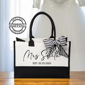 K&ouml;nnte beinhalten: Wei&szlig;e Canvas-Tragetasche mit schwarzen Henkeln und einer schwarz-wei&szlig;en Schleife. Die Tasche tr&auml;gt den Aufdruck "Mrs. Smith EST. 02.25.2025".