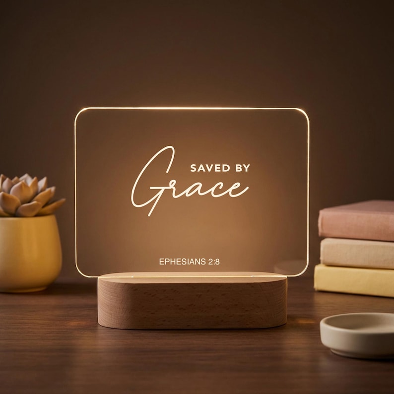 Może przedstawiać: Prostokątna lampka nocna LED z napisem "Saved by Grace" w ozdobnej czcionce. Światło stoi na drewnianej podstawie i wyświetla werset biblijny Efezjan 2:8. Po lewej stronie znajduje się mały sukulent w ż&oacute;łtej doniczce.