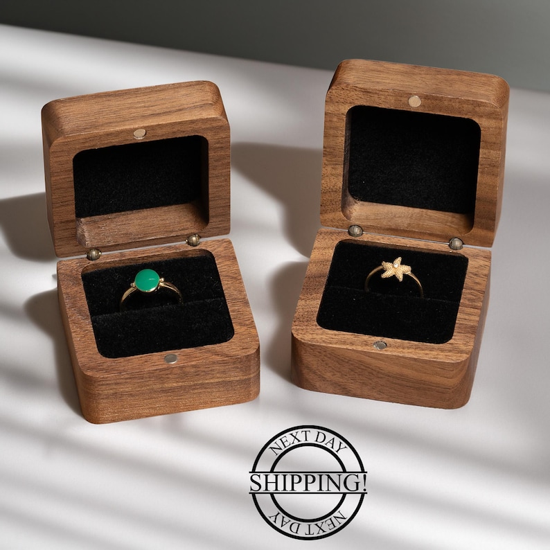 May include: Two open wooden ring boxes with black velvet interiors. One box holds a gold ring with a green stone, the other a gold starfish ring. The boxes are made of light brown wood.