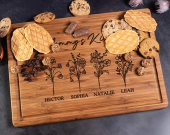 Könnte beinhalten: Ein personalisiertes Bambus-Schneidebrett mit dem Text "Mommy's Kitchen" und vier floralen Designs mit den Namen "Hector", "Sophia", "Natalie" und "Leah", die unter jedes Design graviert sind. Das Brett ist mit Keksen, Waffeln und Schokolade verziert.
