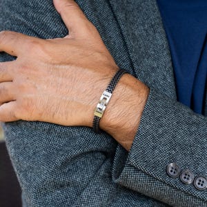May include: A black braided bracelet with a gold clasp. The clasp is engraved with the name "Eddie" and a small diamond. The bracelet is worn on a person's wrist, with a gray blazer in the background.