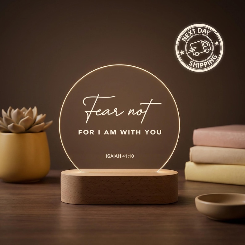 May include: A glowing LED night light with a wooden base. The light features the words "Fear not, for I am with you" in elegant script. A small succulent in a yellow pot and a stack of books are in the background.