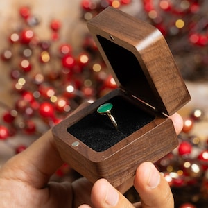 May include: A gold ring with a green oval gemstone is displayed inside a wooden ring box with a black velvet interior. The box is open, revealing the ring. The box is made of dark brown wood.