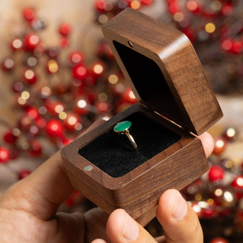 May include: A gold ring with a green oval gemstone is displayed inside a wooden ring box with a black velvet interior. The box is open, revealing the ring. The box is made of dark brown wood.