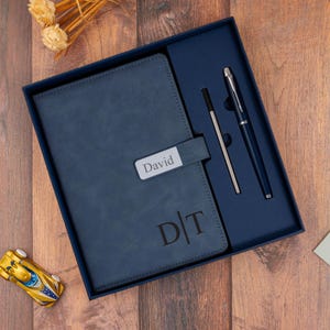 May include: A navy blue gift set in a box, including a notebook with the name "David" and initials "D|T", a pen, and a pen refill. The set is presented on a wooden surface.