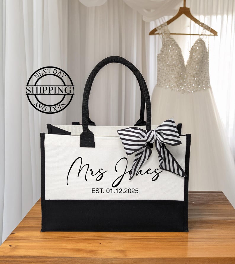 K&ouml;nnte beinhalten: Eine wei&szlig;e und schwarze Tragetasche mit schwarzen Henkeln und einer schwarz-wei&szlig;en Schleife. Die Tasche hat den Text "Mrs. Jones EST. 01.12.2025" auf der Vorderseite.