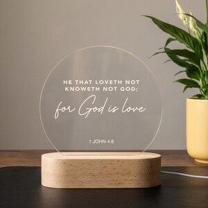 Może przedstawiać: Przezroczysta akrylowa lampka nocna LED z cytatem "HE THAT LOVETH NOT KNOWETH NOT GOD; for God is love" i "1 JOHN 4:8" wydrukowanym na biało. Światło spoczywa na podstawie z naturalnego drewna.