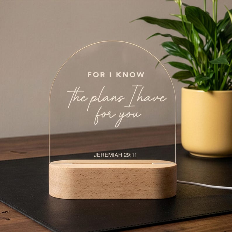 Może przedstawiać: Przezroczysta akrylowa lampka nocna LED z białym napisem "FOR I KNOW the plans I have for you" i "JEREMIAH 29:11". Światło spoczywa na prostokątnej drewnianej podstawie. Roślina w ż&oacute;łtej doniczce znajduje się w tle.