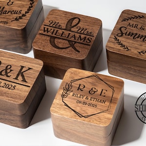 May include: Five wooden ring boxes with engraved names and dates. The boxes are square with hinged lids. The text includes names like "Marcus", "Williams", and "Simmons", along with dates. The boxes are a warm brown color.