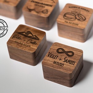 May include: A collection of wooden ring boxes, each with unique engraved details. One box displays an infinity symbol and the names "Violet + Samuel" with the date "06.13.2025". Other boxes feature names and dates.