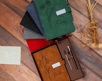 Könnte beinhalten: Eine Sammlung von Notizbüchern in Grün, Blau, Schwarz und Grau sowie ein beiges Notizbuch in einer Geschenkbox mit Stift und Mine. Das grüne Notizbuch zeigt eine Gänseblümchen-Illustration und den Namen Emmy. Das beige Notizbuch zeigt eine Blume und den Namen Sandra.