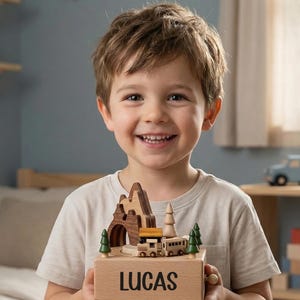 May include: A wooden music box featuring a mountain landscape with a small train, trees, and a tunnel. The base is made of light-colored wood and has the name LUCAS printed in black capital letters on the front side of the rectangular block.