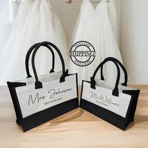 K&ouml;nnte beinhalten: Zwei schwarze und wei&szlig;e Tragetaschen mit Henkeln. Die Taschen haben einen wei&szlig;en Canvas-Bereich mit schwarzem Text, der "Mrs. Johnson EST. 04.26.2024" und "Mrs. Williams EST. 08.18.2024" lautet.