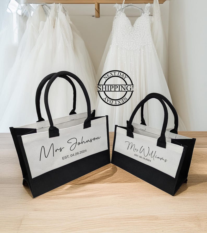 K&ouml;nnte beinhalten: Zwei schwarze und wei&szlig;e Tragetaschen mit Henkeln. Die Taschen haben einen wei&szlig;en Canvas-Bereich mit schwarzem Text, der "Mrs. Johnson EST. 04.26.2024" und "Mrs. Williams EST. 08.18.2024" lautet.