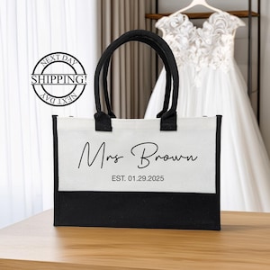 K&ouml;nnte beinhalten: Wei&szlig;e Canvas-Tragetasche mit schwarzen Henkeln und schwarzem Boden. Die Tasche tr&auml;gt den Aufdruck "Mrs. Brown EST. 01.29.2025" auf der Vorderseite.