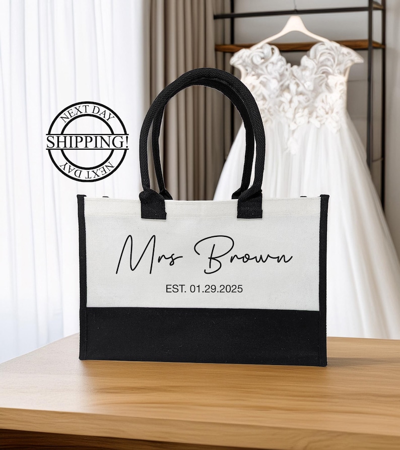 K&ouml;nnte beinhalten: Wei&szlig;e Canvas-Tragetasche mit schwarzen Henkeln und schwarzem Boden. Die Tasche tr&auml;gt den Aufdruck "Mrs. Brown EST. 01.29.2025" auf der Vorderseite.