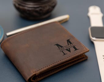 Könnte beinhalten: Ein braunes Leder-Portemonnaie mit einem dunkel gravierten Monogramm-Design auf der Vorderseite. Die Gravur zeigt einen großen Buchstaben M, wobei der Name MICHAEL in einer klaren Serifenschrift horizontal durch die Mitte verläuft.