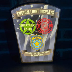 May include: A trapezoidal light display featuring a black and white American flag background. It displays the text CUSTOM LIGHT DISPLAYS, a green military star, a red fire department emblem, a blue police badge, and Your Badge Your Patch Your Design Any Shape Possible.