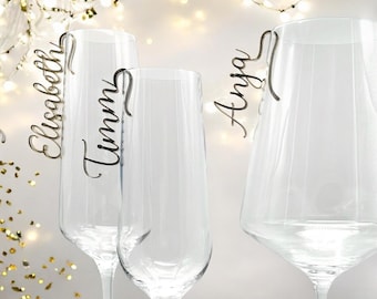 May include: Three clear glass champagne flutes with personalized name tags. The tags are made of wood and are attached to the glass with a wire loop. The names on the tags are "Elisabeth", "Timm", and "Anja".