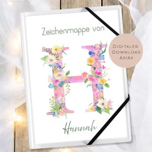 May include: A white art portfolio with a pink floral letter "H" and the name "Hannah" in green script. The letter is adorned with colorful flowers. The portfolio has a black elastic band closure and the text "DIGITALER DOWNLOAD A3/A4".