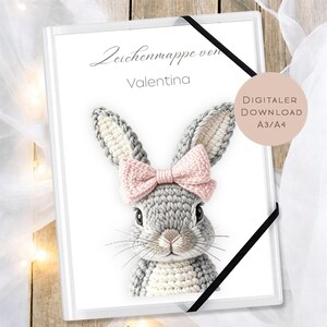May include: A white binder with a black elastic closure featuring a detailed illustration of a gray and white bunny with a pink bow. The text "Zeichenmappe von Valentina" is at the top. A digital download is available.