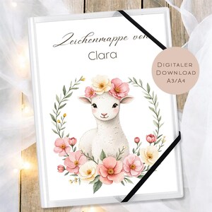 May include: White art portfolio with the text "Zeichenmappe von Clara" and a watercolor illustration of a lamb wearing a floral crown. The lamb is surrounded by a wreath of pink and cream flowers and green leaves. A black elastic band secures the portfolio.