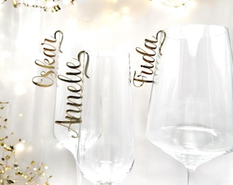 May include: Three clear glass wine glasses with gold-colored wooden name tags attached to the stems. The names on the tags are "Oskar", "Annelie", and "Frida".