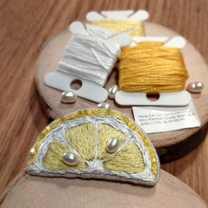 May include: A yellow and white embroidered lemon slice with three white pearl beads. The lemon slice is on a wooden surface with two spools of thread, one white and one yellow.