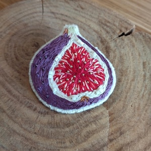 May include: A hand-stitched embroidered fig brooch with a purple exterior and a red interior. The fig is stitched with white thread and has a white outline. The fig is on a wooden surface.