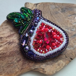 May include: A handcrafted brooch in the shape of a fig, featuring a vibrant design. The brooch is adorned with red, purple, green, and white beads, creating a detailed and eye-catching accessory. The fig's interior is filled with red beads, and the exterior is outlined with purple beads.