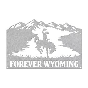 May include: A silver-colored metal wall sign featuring a cutout silhouette of a bucking horse and rider against a mountain range backdrop. The text FOREVER WYOMING is cut out at the bottom. The piece has small circular holes on the left and right sides.