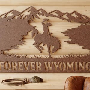 May include: A brown metal wall art piece featuring a silhouette of a cowboy riding a bucking horse against a mountain range, with the text FOREVER WYOMING below. Included are a coiled rope and a metal belt buckle with a bucking horse design.