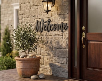 May include: A black metal Welcome sign mounted on a stone wall next to a dark wood door. A small olive tree sits in a terracotta pot on a wooden porch, accompanied by a black outdoor wall lantern and a tan doormat with a letter H monogram.