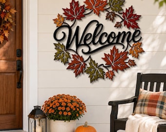 May include: A circular wall sign featuring the word Welcome in black cursive script, surrounded by a wreath of metallic maple leaves in shades of red, orange, yellow, and green. The sign is mounted on a white horizontal paneled wall.