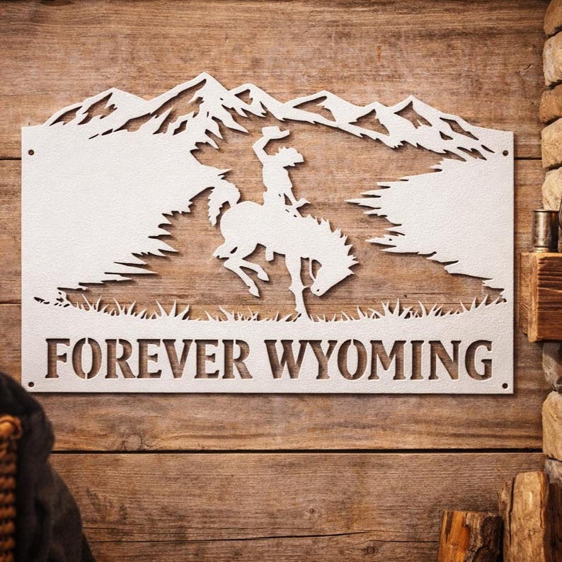 May include: A white rectangular metal wall sign featuring a laser-cut silhouette of a cowboy riding a bucking horse against a mountain range backdrop. The text FOREVER WYOMING is cut out in a bold serif font along the bottom edge of the sign.