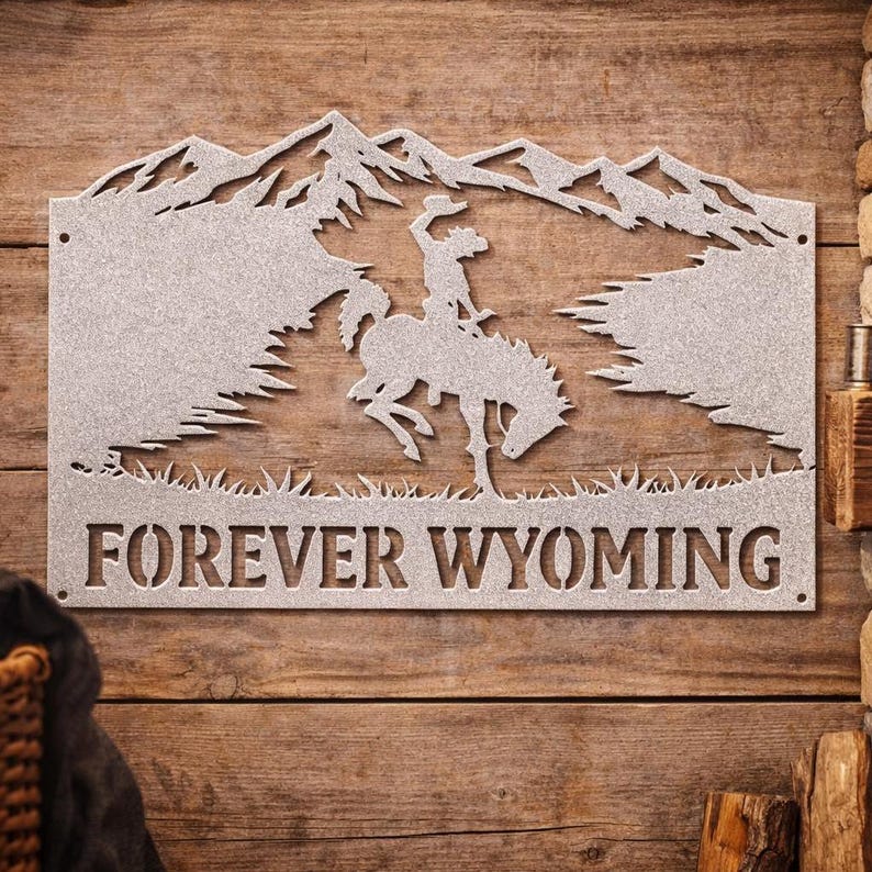 May include: A silver-colored metal wall sign featuring a cutout silhouette of a person riding a bucking horse in front of mountain peaks. The text FOREVER WYOMING is cut out at the bottom of the rectangular piece, which has small mounting holes.
