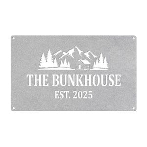 May include: A rectangular, silver-colored metal sign with the words "THE BUNKHOUSE" and "EST. 2025" cut out in a bold font. Above the text is a mountain and cabin scene, also cut out. Four small holes are in each corner.