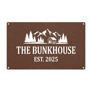 May include: A rectangular brown metal sign with the words "THE BUNKHOUSE EST. 2025" in white. The sign features a white graphic of mountains, trees, and a cabin. Four small holes are in each corner.