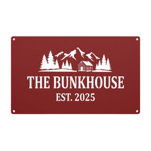 May include: A rectangular, burgundy metal sign with white text and a mountain and cabin design. The text reads "THE BUNKHOUSE EST. 2025". The sign has four holes, one in each corner.