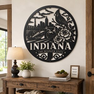 Może przedstawiać: Czarne metalowe dzieło sztuki na ścianę w okrągłym kształcie z napisem "INDIANA" i "THE HOOSIER STATE". Grafika zawiera samoch&oacute;d wyścigowy, panoramę miasta, ptaka i elementy kwiatowe. Dzieło sztuki jest eksponowane na białej ścianie.