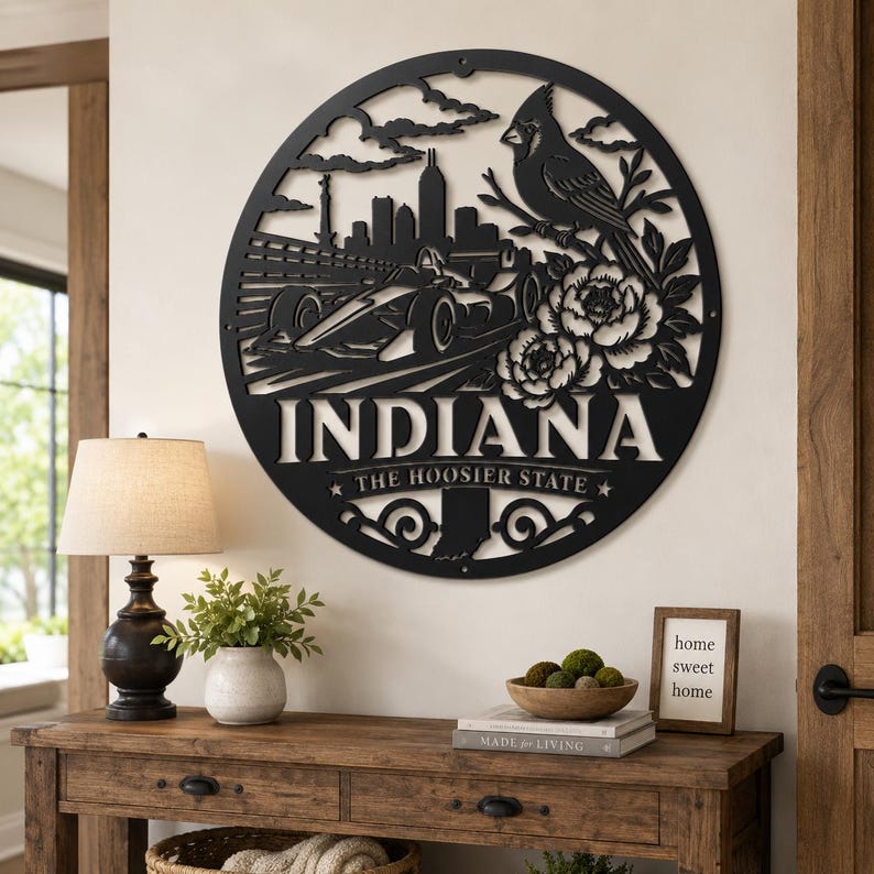 Może przedstawiać: Czarne metalowe dzieło sztuki na ścianę w okrągłym kształcie z napisem "INDIANA" i "THE HOOSIER STATE". Grafika zawiera samoch&oacute;d wyścigowy, panoramę miasta, ptaka i elementy kwiatowe. Dzieło sztuki jest eksponowane na białej ścianie.