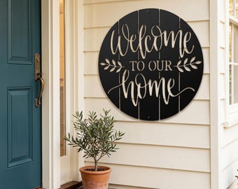 May include: A circular black metal wall sign featuring the cutout text "Welcome TO OUR home" in a script and sans-serif font combination. The design includes decorative leaf motifs on both sides of the text, with vertical slats visible.