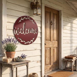 May include: A circular dark red metal wall sign featuring the word welcome in a cursive script, surrounded by a laser-cut leafy branch border. The sign is mounted on a light-colored wooden wall next to a wooden door and a potted lavender plant.