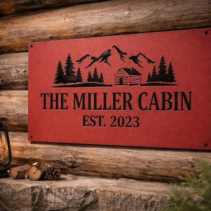 May include: A red metal sign with a mountain and cabin silhouette design. The sign reads "THE MILLER CABIN EST. 2023" in black lettering. The sign is mounted on a wooden log wall.