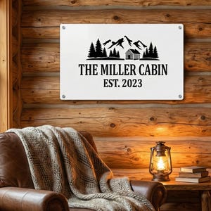 May include: A white metal sign with the text "THE MILLER CABIN EST. 2023" and a black graphic of mountains, trees, and a cabin. The sign is mounted on a wooden wall. A leather armchair with a blanket, a lantern, and books are in the foreground.