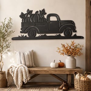 May include: A matte black metal wall art piece shaped like a vintage pickup truck carrying three pumpkins in its bed, accented with leaf motifs. The design features a flat base with grass silhouettes and two mounting holes on the ends.